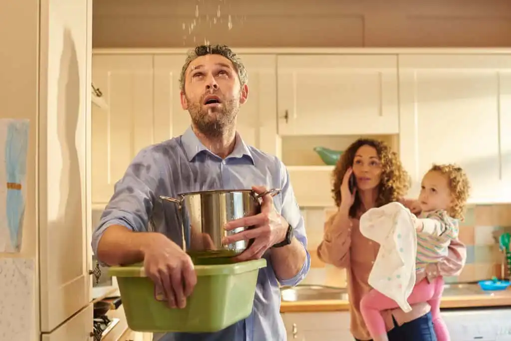 A man stands in a kitchen holding a pot and a basin to catch water dripping from the ceiling, while a concerned woman holding a child watches in the background.