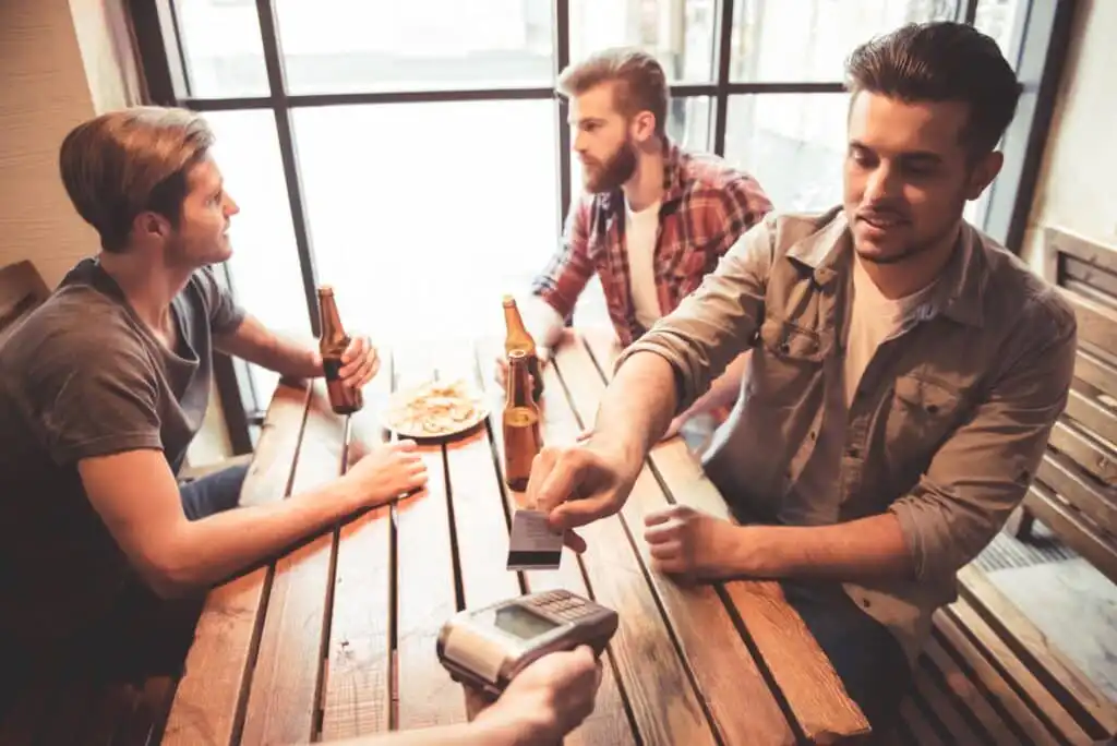 Three men sit at a wooden table in a cafe, two holding beer bottles, with food in the center. One man extends a card to pay with a portable card reader held by an unseen person.