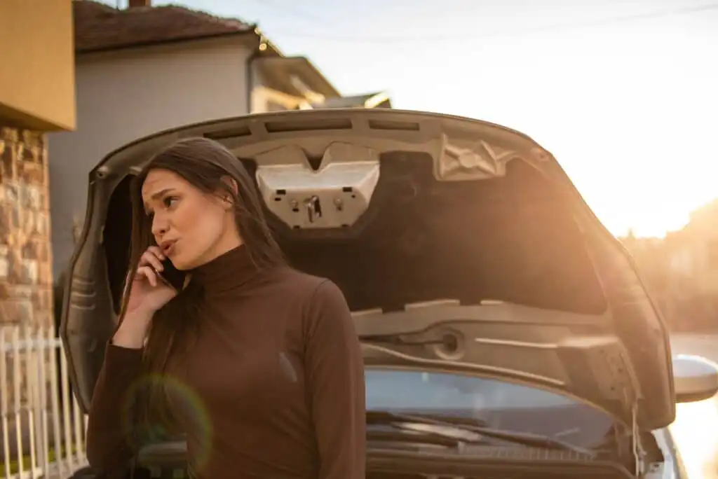 A woman stands in front of a car with its hood open, talking on her phone, appearing concerned. The scene is outdoors with sunlight in the background.
