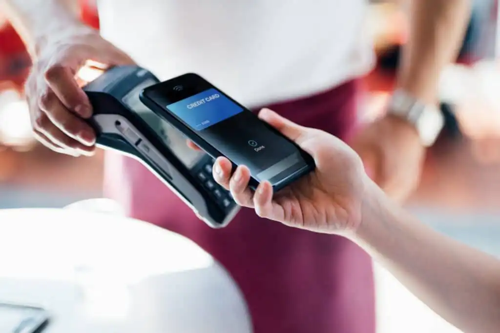 A person holds a smartphone near a card reader, making a contactless payment using a mobile wallet app, while another person holds the payment terminal.