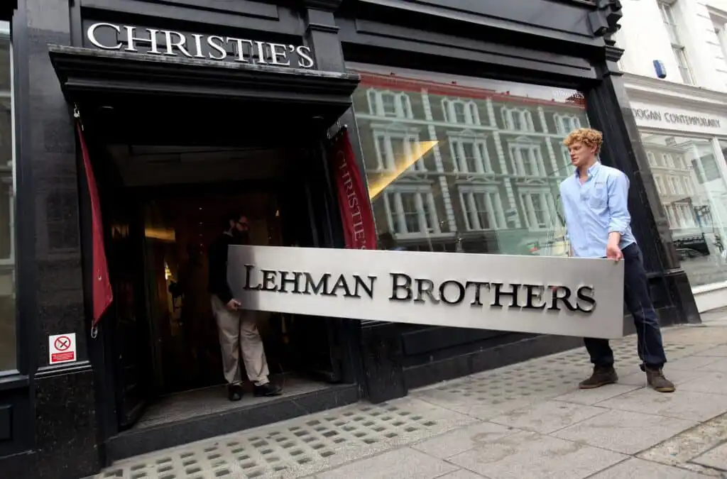Two men carry a large Lehman Brothers sign out of the entrance to Christies auction house, with the building’s black facade and a Christies sign visible above the door.