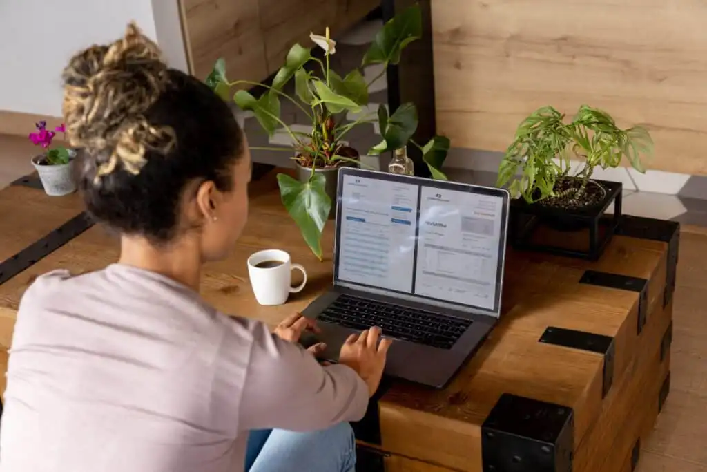 A woman sits at a wooden coffee table, working on a laptop with two open tabs. A mug, several potted plants, and a mirror are also on the table. She is focused on her screen.