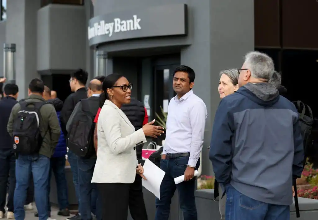 A group of people stand in line outside a Silicon Valley Bank branch. Some are talking while others wait, holding papers and bags, with the bank’s entrance sign visible in the background.