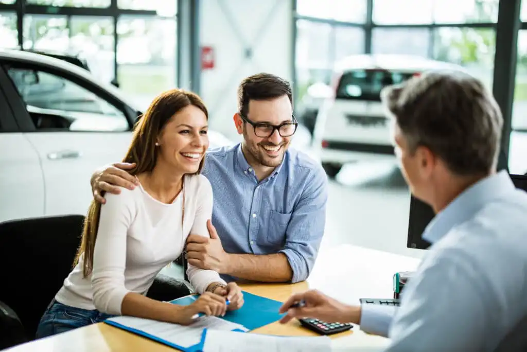 A smiling couple sits at a desk across from a professional, discussing documents in a bright office with cars visible in the background. The atmosphere appears friendly and collaborative.