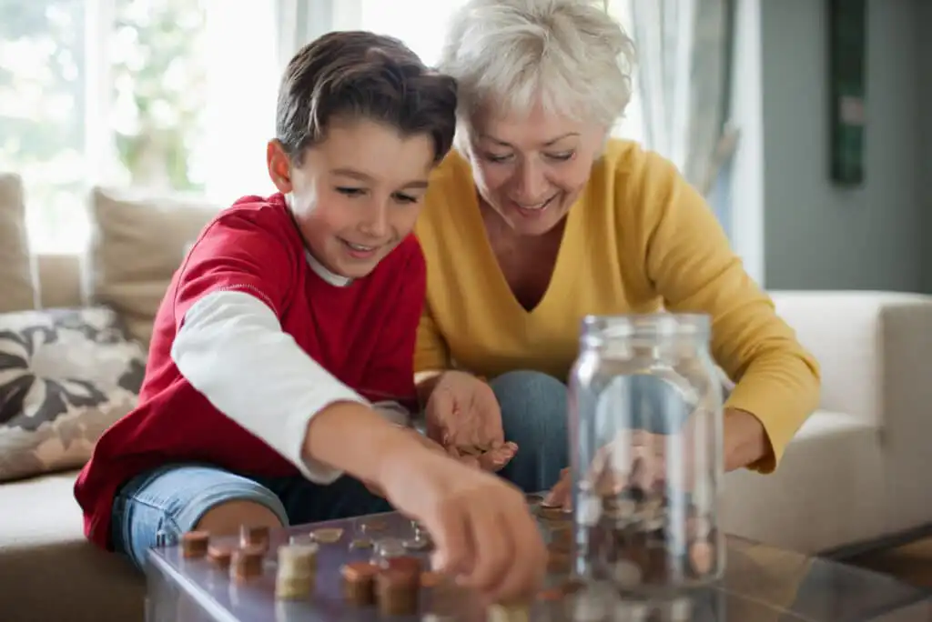 A smiling elderly woman and a young boy sit together on a couch, sorting and stacking coins on a glass table. A large jar filled with coins sits in front of them, creating a cheerful, bonding moment.