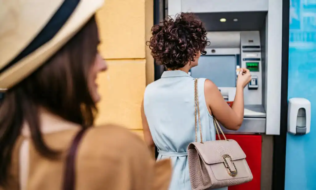 A woman with curly hair and a beige handbag uses an outdoor ATM, while another woman in a hat stands blurred in the foreground, watching her.