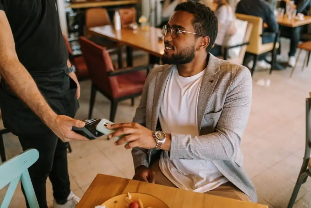 A man in a light gray blazer is sitting at a restaurant table, smiling and paying for his meal with a smartphone using contactless payment held by a server.