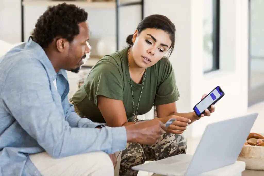 A woman shows a smartphone screen to a man while they sit together at a table with a laptop. They appear to be having a serious discussion in a casual indoor setting.