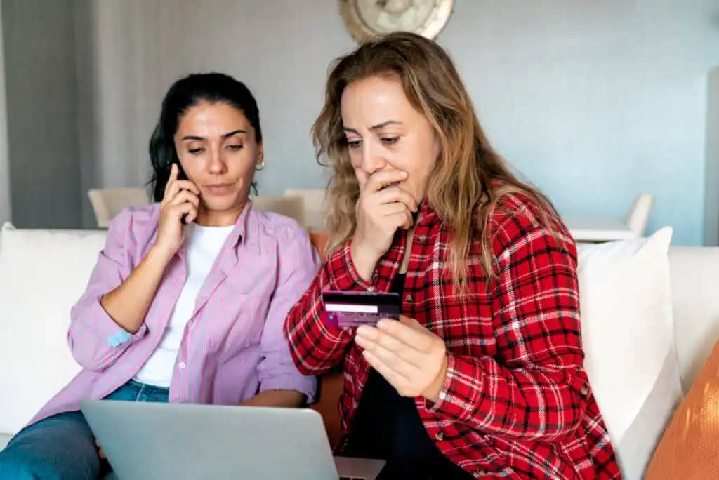 Two worried women sit on a couch with a laptop. One is on the phone, while the other holds a credit card and covers her mouth, suggesting concern or stress about an online transaction.