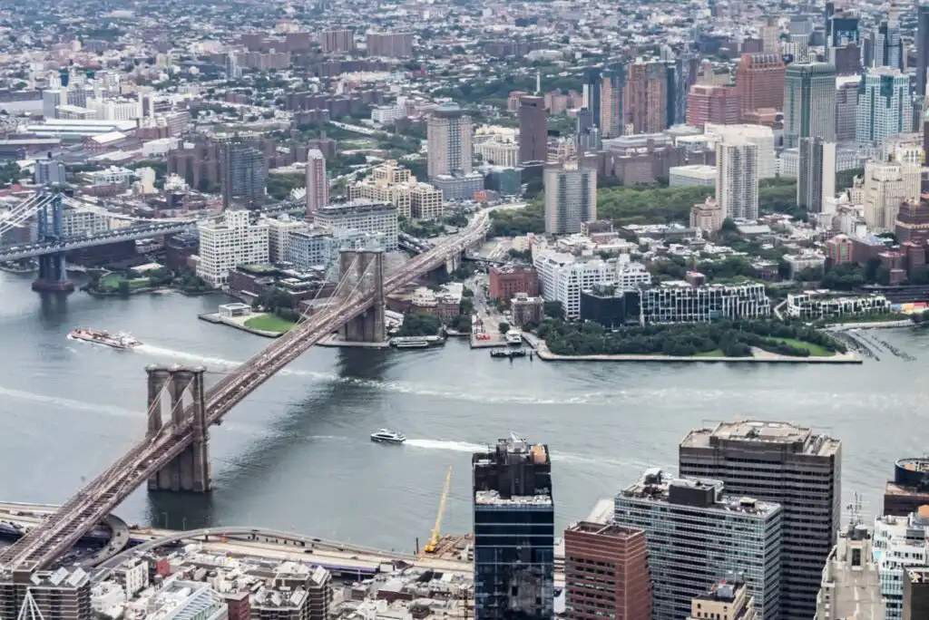 Aerial view of the Brooklyn Bridge spanning the East River, connecting Manhattan and Brooklyn with city buildings, parks, and boats visible on the water below.