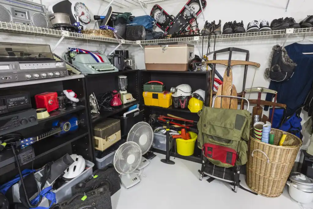 A cluttered storage room with shelves holding various items like electronics, helmets, boxes, fans, tools, bags, and winter gear. Sports equipment and baskets are also visible, all organized in a small space.