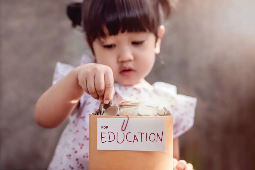 A young girl places coins into a container labeled For Education, symbolizing saving money for educational purposes. The background is softly blurred, highlighting her focused expression.