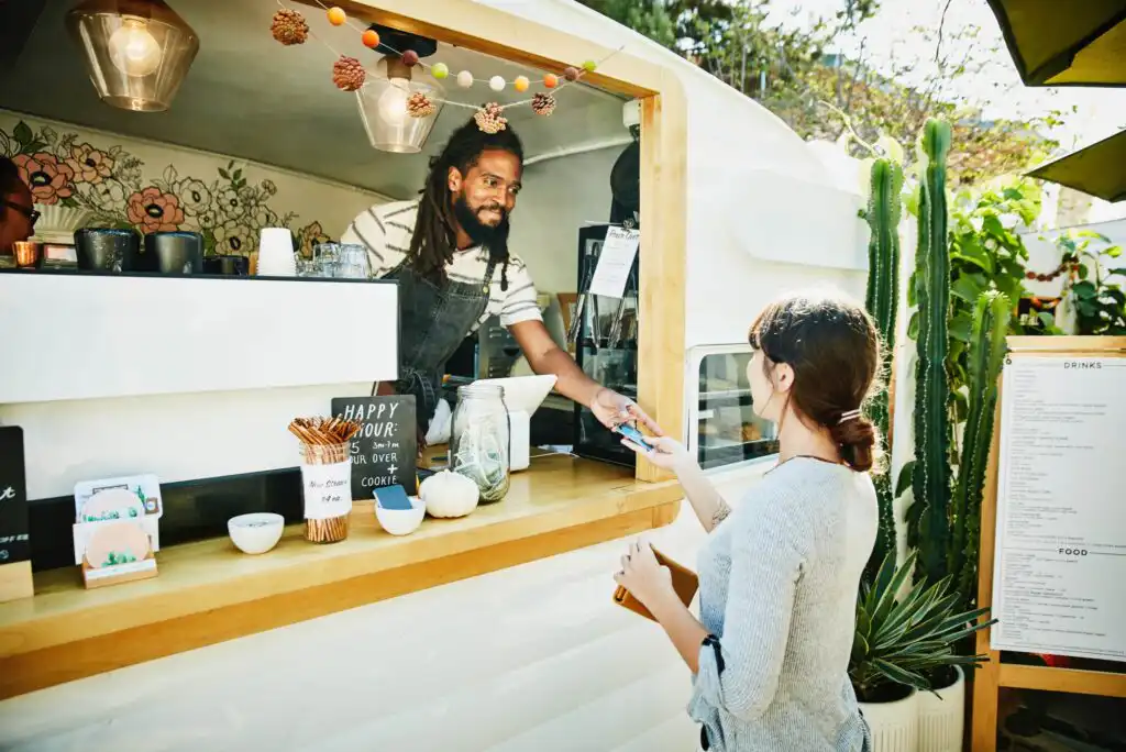 A man in an apron serves a woman at a food truck window decorated with lights and small pumpkins. The woman pays with a card while holding a drink, and the man smiles at her from inside the truck.