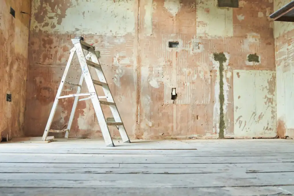 A metal ladder stands on a wooden floor in front of a stripped, unfinished wall with exposed plaster and patches, suggesting a room under renovation or construction.