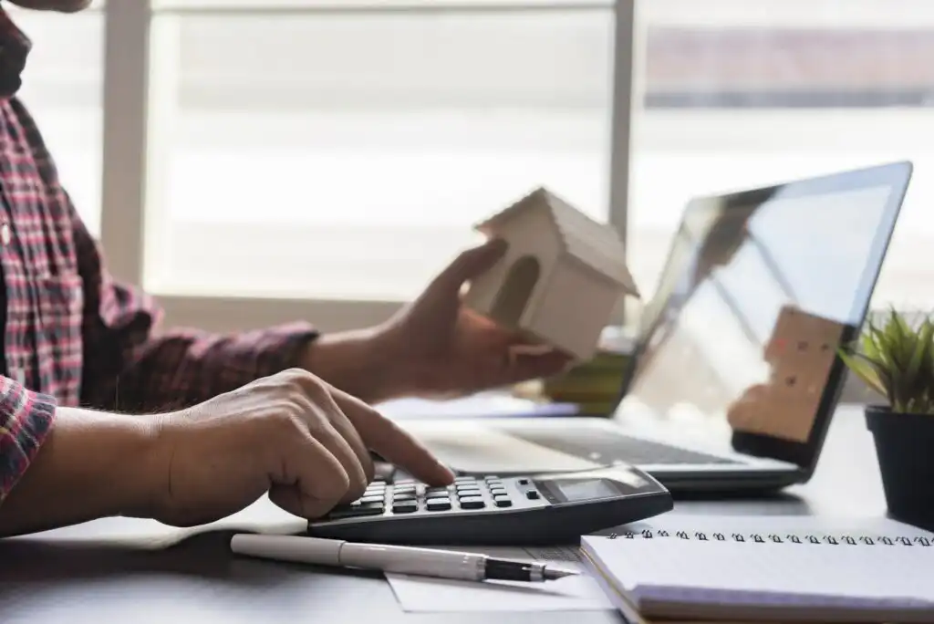 A person sits at a desk using a calculator with one hand and holding a small model house with the other. A laptop, notepad, pen, and small potted plant are on the desk by a window.