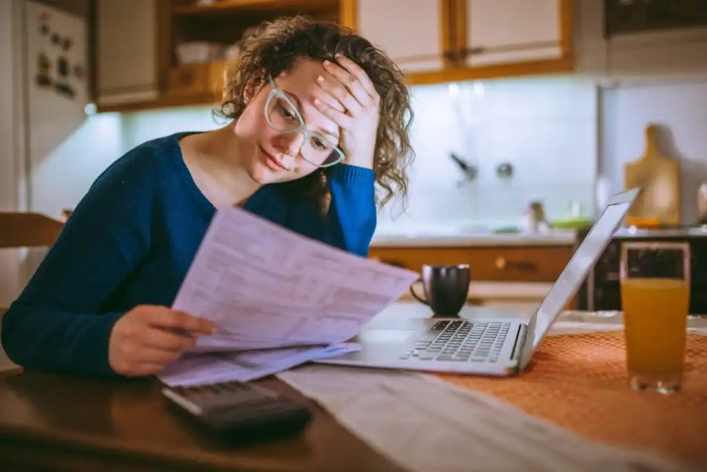 A woman with glasses looks stressed while reviewing papers at a kitchen table with a laptop, calculator, coffee mug, and a glass of orange juice.