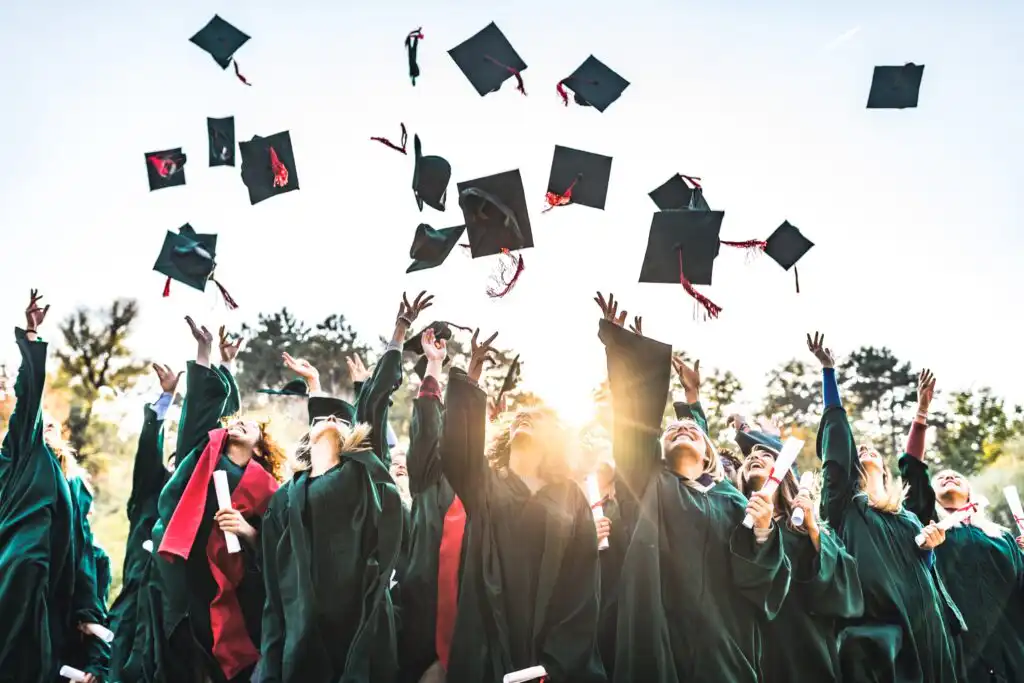 A group of graduates in caps and gowns celebrate outdoors, tossing their graduation caps into the air against a bright sky.