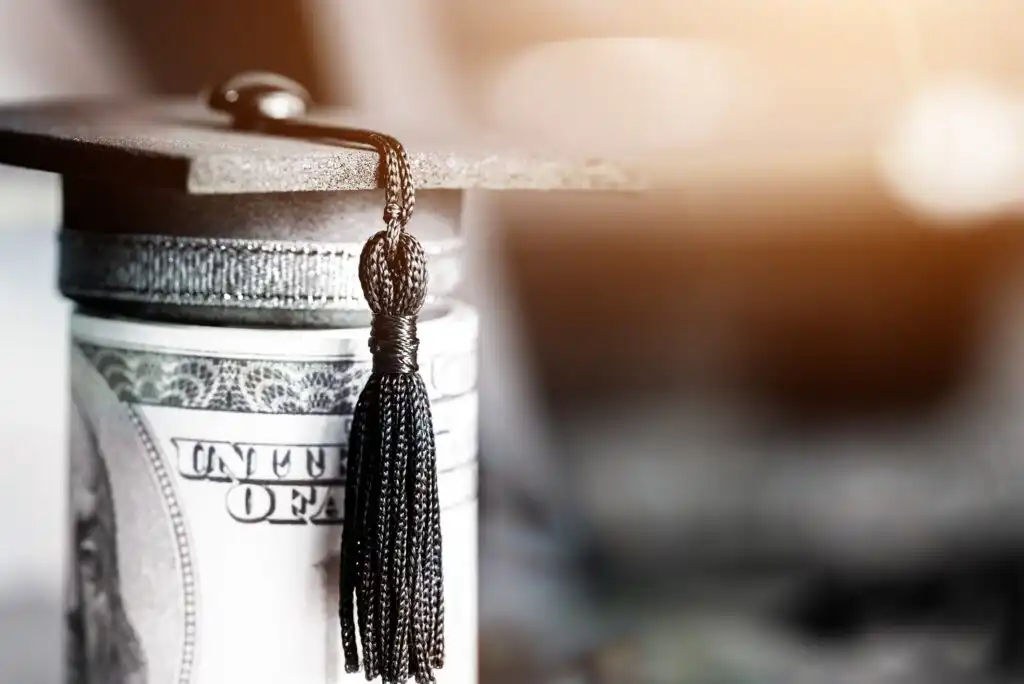 A rolled-up U.S. hundred-dollar bill wrapped with a black graduation cap and tassel, symbolizing student loans, education costs, or financial aid for higher education.