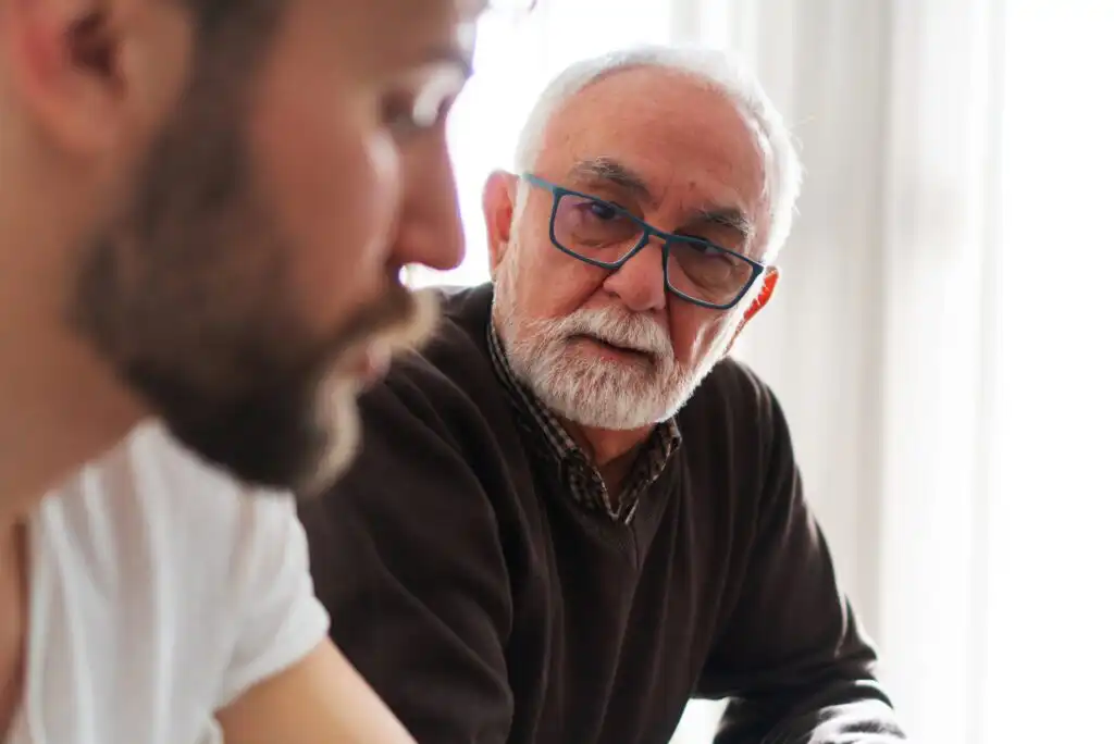 An older man with white hair, a beard, and glasses looks intently at a younger man with a beard, who is out of focus in the foreground. They appear to be having a serious conversation indoors.