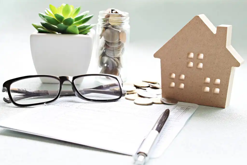A pair of eyeglasses, a pen, a document, a small potted succulent, a jar of coins, and a wooden house model are arranged on a light surface, symbolizing financial planning for home ownership.