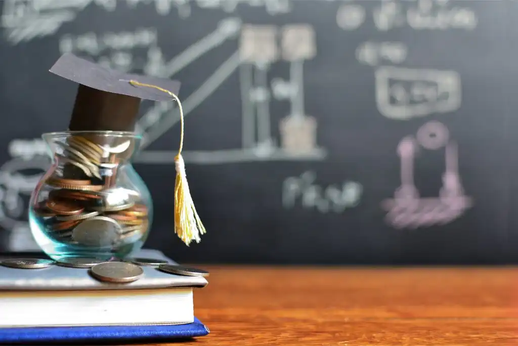 A glass jar filled with coins sits on top of books, with a graduation cap on the jar. A chalkboard with diagrams and formulas is blurred in the background.