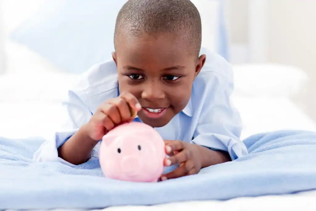 A young boy smiling as he puts a coin into a pink piggy bank while lying on a bed covered with a blue blanket.