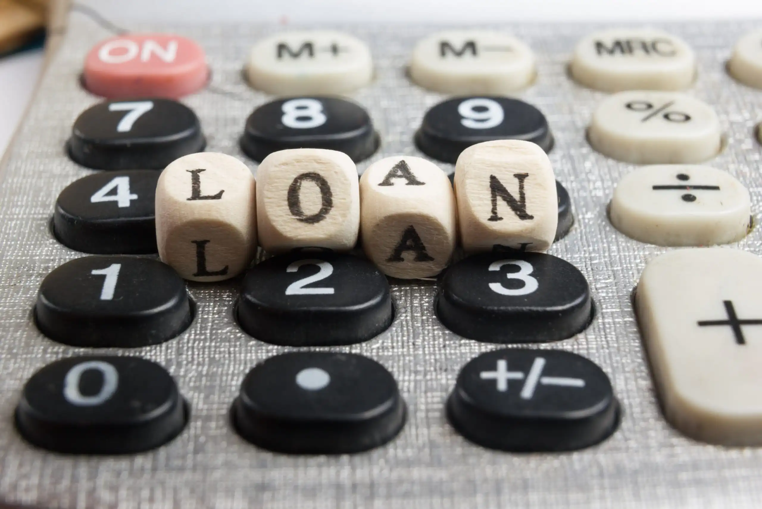 Wooden letter blocks spelling LOAN rest on the keys of a calculator, symbolizing financial calculations or considerations related to loans.