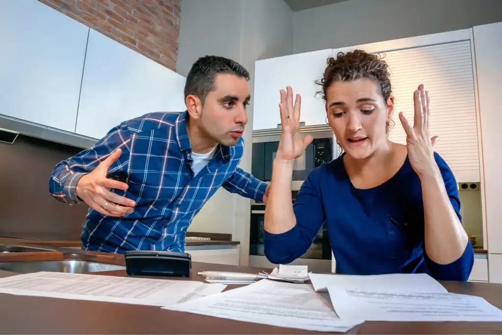 A man and woman argue in a kitchen, surrounded by papers and a calculator. The woman looks frustrated, raising her hands, while the man gestures with concern, leaning toward her over the counter.