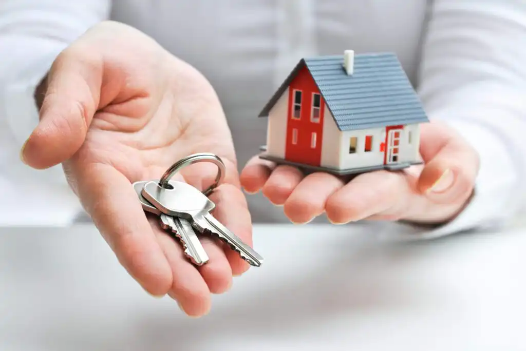 A person holds a set of house keys in one hand and a small model of a house in the other, symbolizing buying, selling, or renting a home.