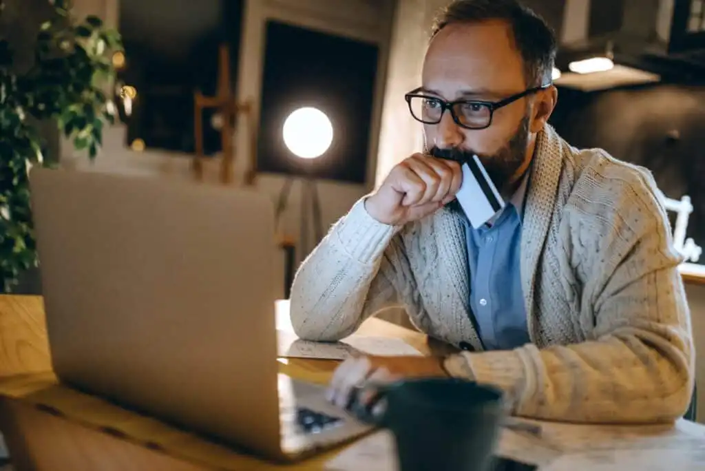 A man wearing glasses and a sweater sits at a table, looking thoughtfully at a laptop while holding a credit card to his mouth. Papers and a cup are on the table, with a lamp glowing in the background.