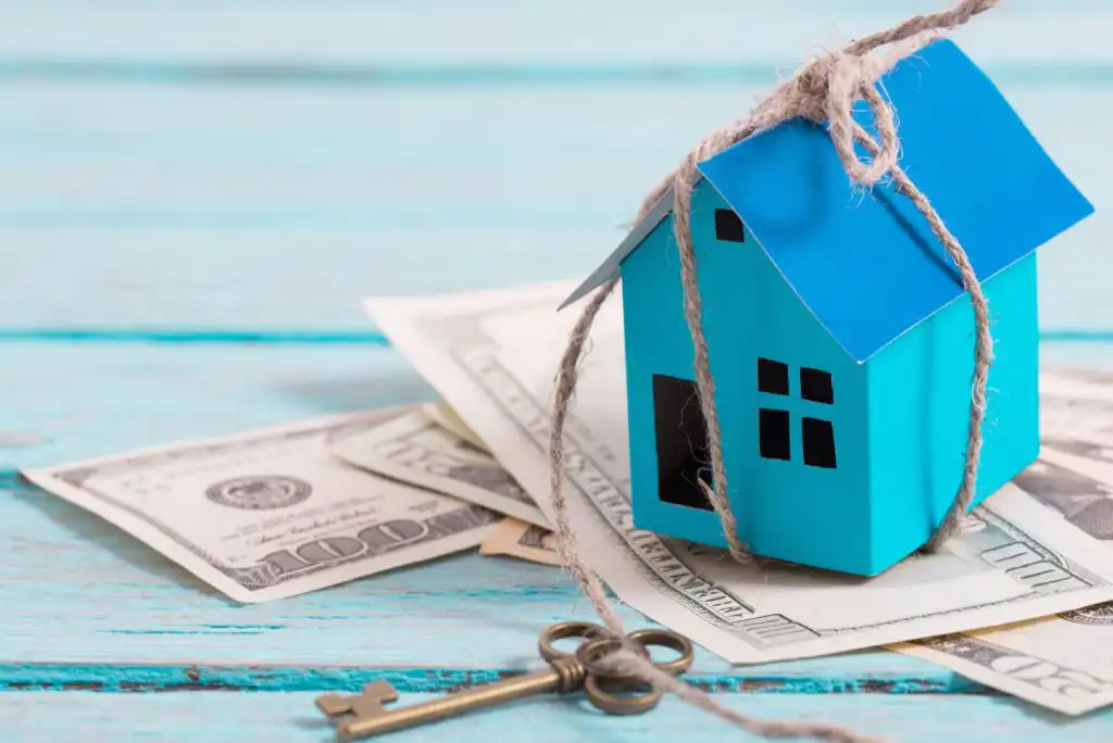 A small blue paper house tied with string sits on scattered US dollar bills next to an old-fashioned key, all on a light blue wooden surface.