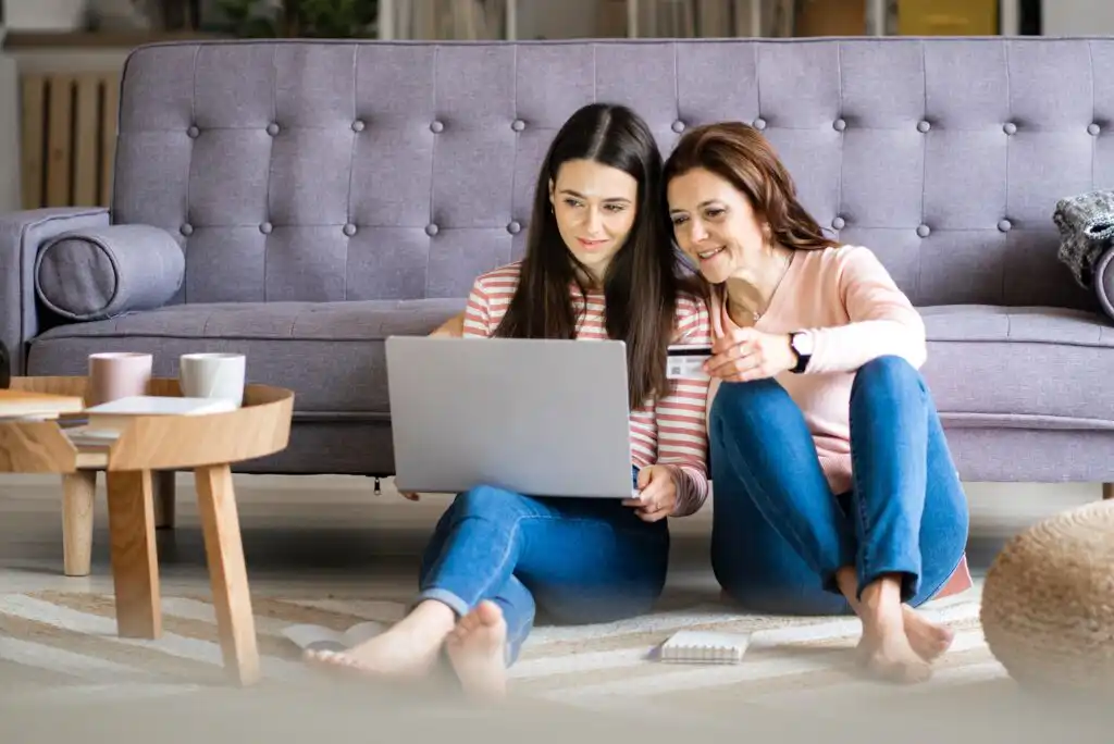 Two women sitting on the floor in front of a sofa, smiling and looking at a laptop together. One is holding a credit card. There is a coffee table, notebook, and pen nearby.