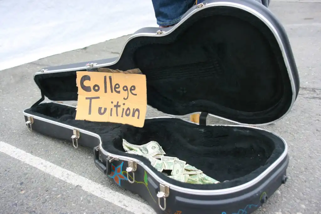 An open guitar case with a cardboard sign reading “College Tuition” inside, containing several dollar bills, sits on the pavement, suggesting street performing to raise money for education.