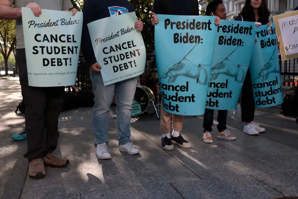 Four people stand holding blue signs that read “President Biden: CANCEL STUDENT DEBT!” during a protest outdoors; their faces are not visible in the photo.