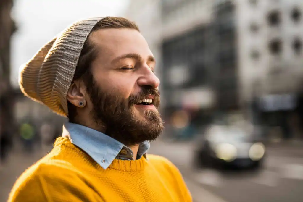 A bearded person wearing a gray knit hat, yellow sweater, and blue shirt smiles with eyes closed while standing outdoors on a city street with blurred buildings and cars in the background.
