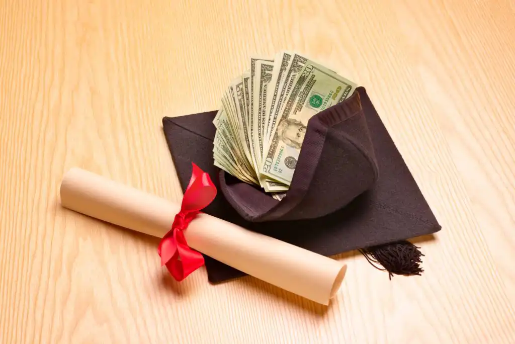 A graduation cap filled with U.S. dollar bills sits next to a diploma tied with a red ribbon on a wooden surface.