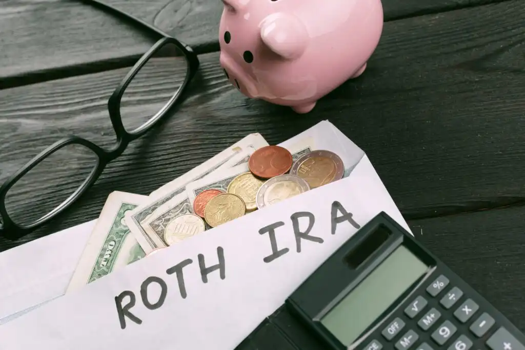 A pair of glasses, a pink piggy bank, and a calculator sit on a table beside an envelope labeled ROTH IRA, filled with various paper bills and coins.