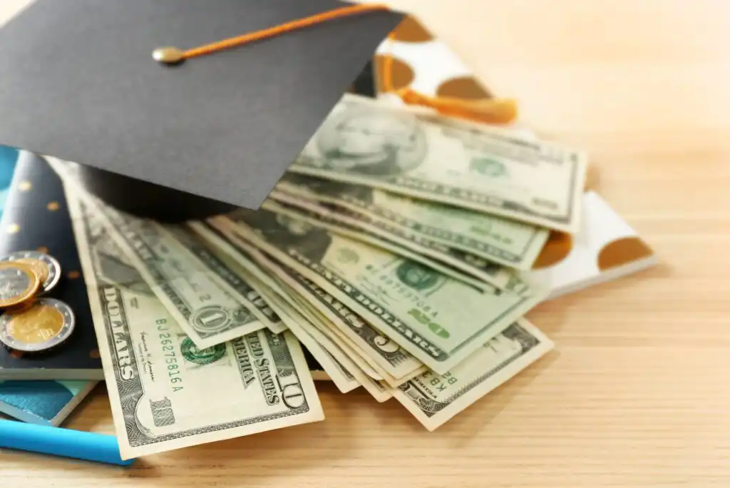 A graduation cap resting on a stack of U.S. dollar bills, coins, and a polka-dotted envelope on a wooden surface, symbolizing the cost or funding of education.