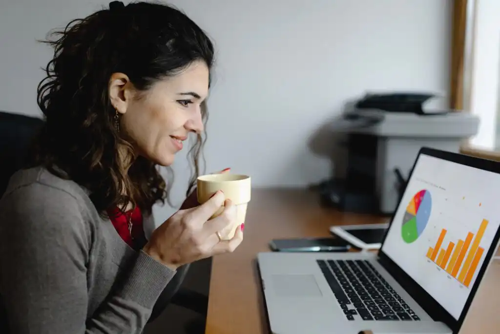 A woman sits at a desk holding a mug and smiling while looking at a laptop displaying colorful charts and graphs. A smartphone and office supplies are on the desk beside her.