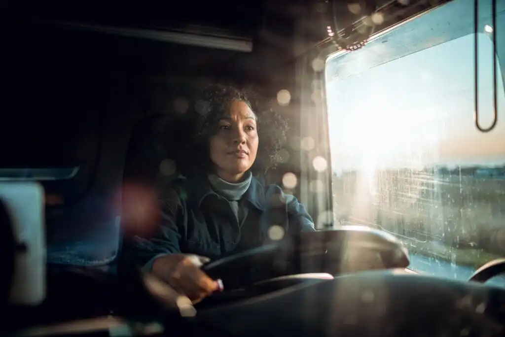 A woman with curly hair drives a vehicle, looking ahead with a focused expression. Sunlight streams through the side window, illuminating her face and the dashboard. The photo is taken from inside the vehicle.