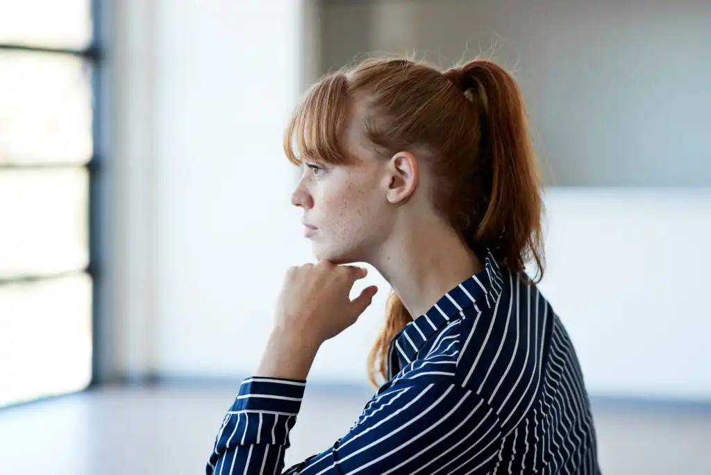 A young woman with red hair in a ponytail, wearing a navy blue and white striped shirt, sits indoors looking thoughtfully to the side with her chin resting on her hand.