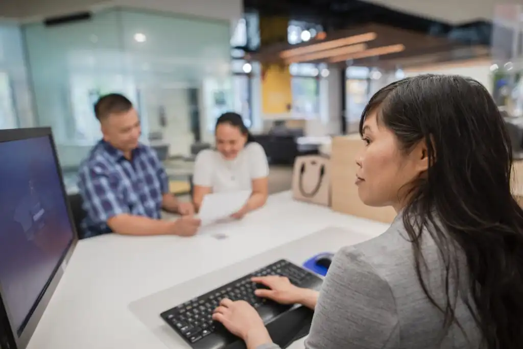A woman in a gray blazer works at a computer, while a man and woman sit across the desk reviewing paperwork together in a modern office setting.