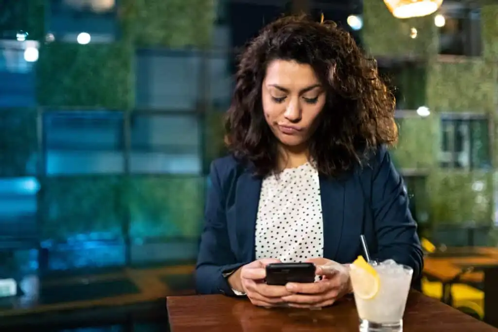A woman with curly hair in business attire sits at a table in a modern bar, looking down at her phone with a serious expression. A cocktail with a lemon slice and straw is on the table in front of her.