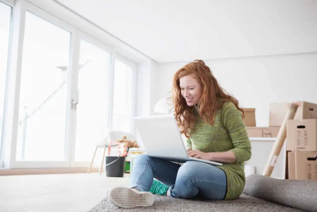 A woman with red hair sits cross-legged on a carpeted floor, smiling and using a laptop. She is in a bright room with large windows and moving boxes in the background.