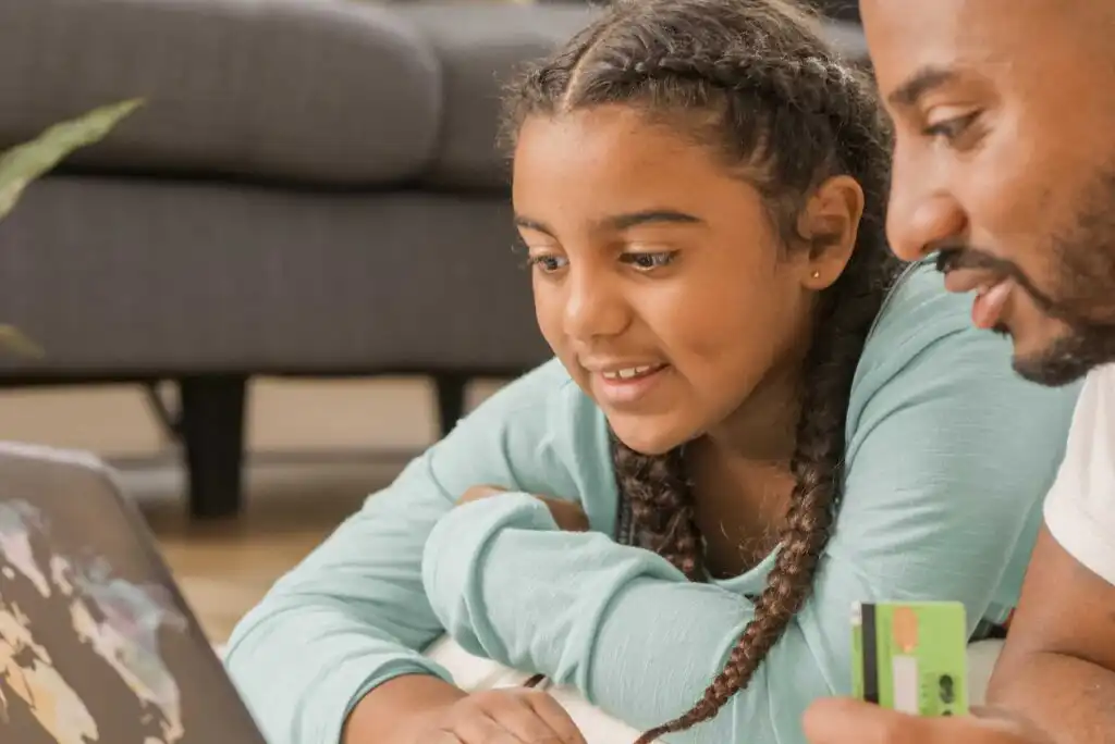 A young girl with braids and a man smile while looking at a laptop screen. The man holds a green credit card, suggesting they are online shopping or learning together at home.