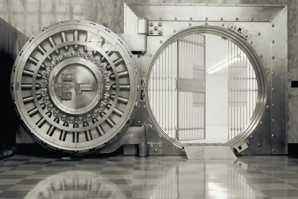 A large metal bank vault door stands open, revealing a bright, empty room with barred metal gates inside. The polished floor reflects the door and surroundings.