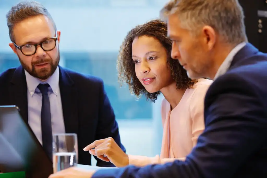 Three professionally dressed people sit at a table, focused on a laptop screen. One woman is pointing at the display while the two men attentively watch, suggesting a collaborative business meeting.