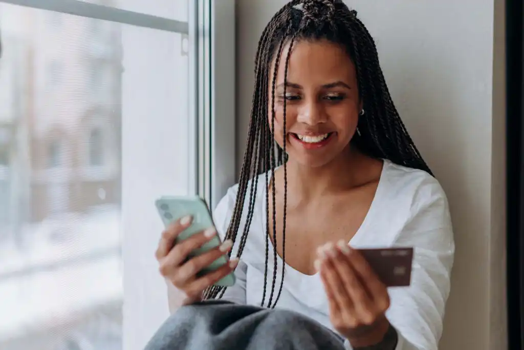 A smiling woman with long braids sits by a window holding a smartphone in one hand and a credit card in the other, appearing to make an online purchase.