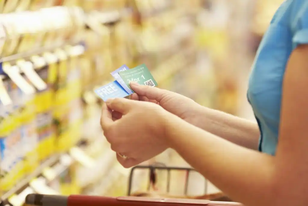 A person in a blue shirt holding coupons while standing next to a grocery store shelf, with a shopping cart visible in the foreground.