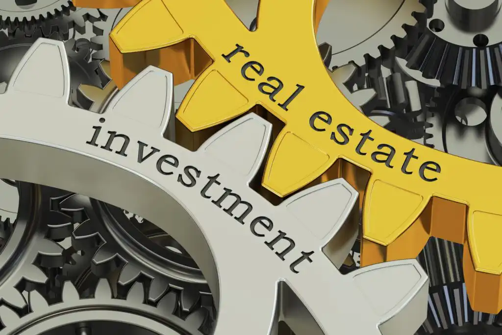 Close-up of interlocking metal gears with the words “real estate” on a gold gear and “investment” on a silver gear, symbolizing the connection between real estate and investment.